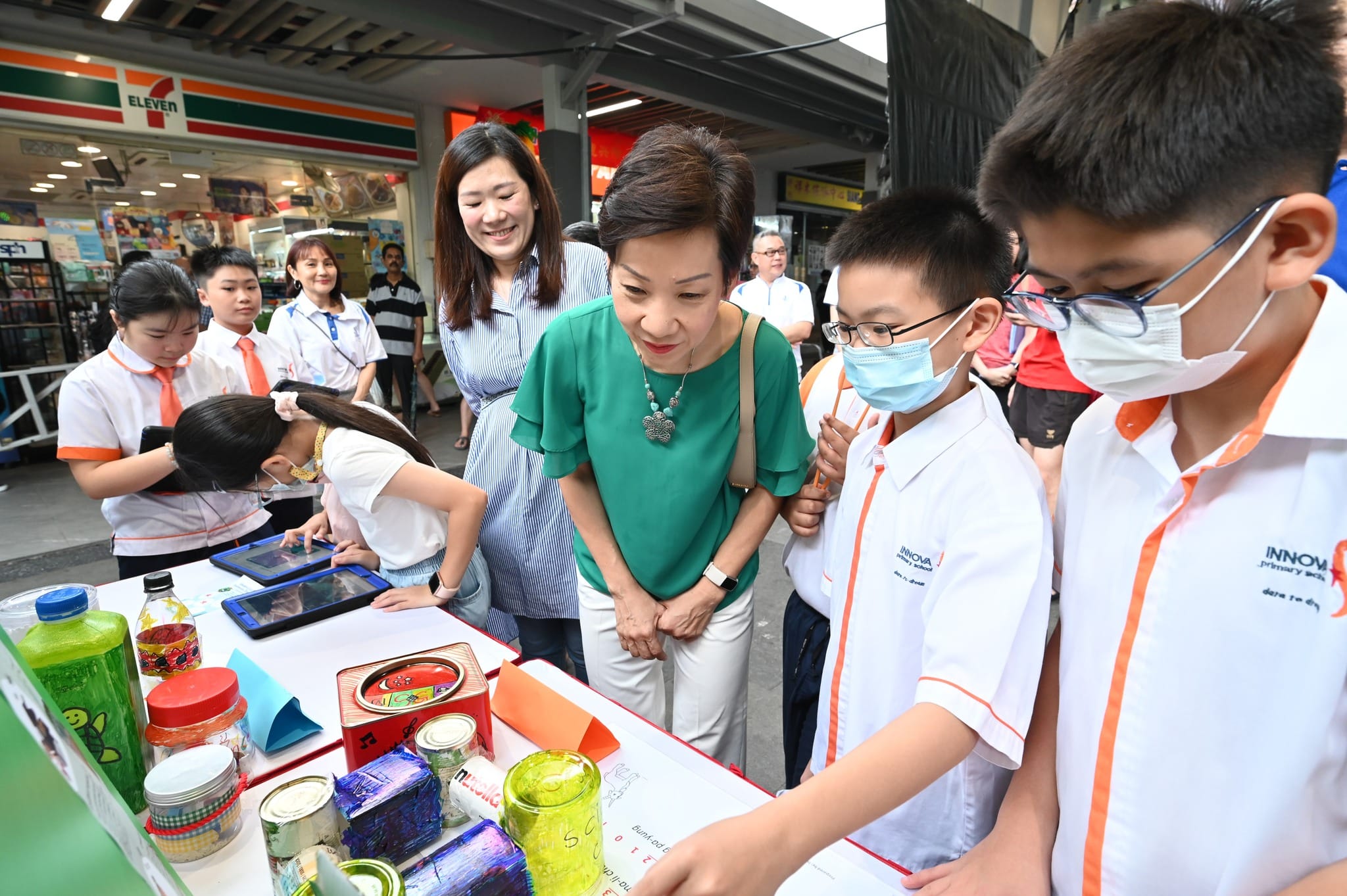 Adults and masked primary school students in white uniforms gather around an outdoor display table near a 7-Eleven store.